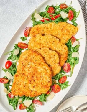 Chicken milanese cutlets on a bed of arugula salad on a serving platter. Next to this is a plate with 2 forks.