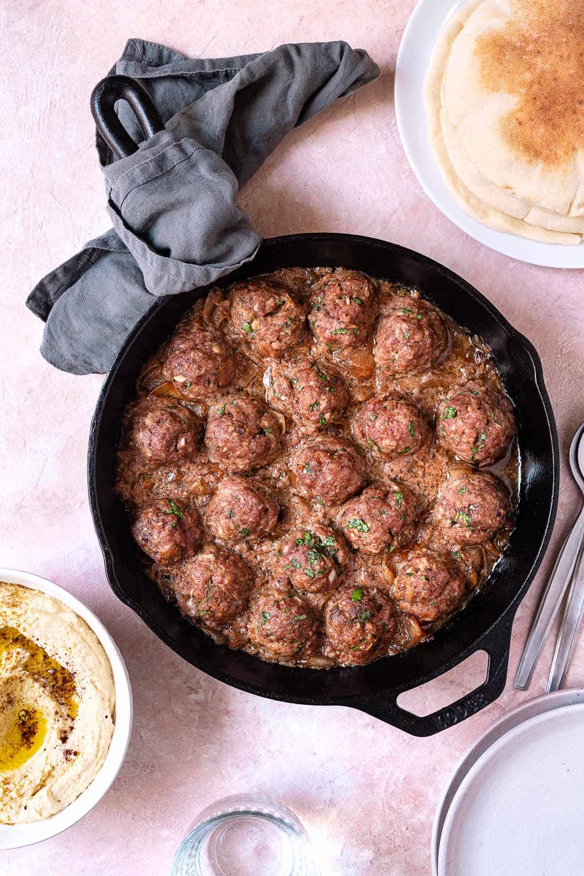 juicy lamb meatballs and an onion sauce in a cast iron skillet next to a side of hummus and pita bread.