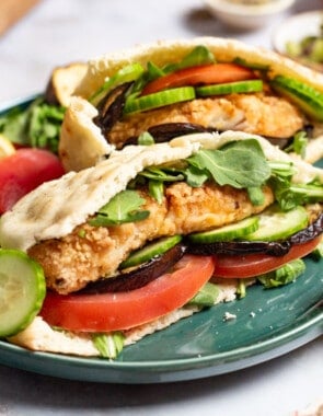 A close up of two fried fish sandwiches on a plate with sides of roasted eggplant slices tomato, persian cucumber slices and a lemon wedge. In the background is a small bowl of olives.