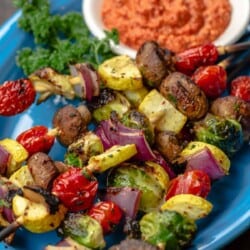A blue plate with several grilled vegetable kabobs and a dish of romesco sauce