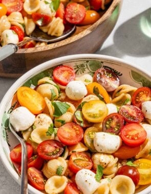A serving of caprese salad in a bowl with a fork next to the rest of the salad in a serving bowl with a spoon.