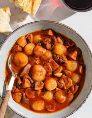 A serving of mushroom stew in a bowl with a spoon next to pieces of crusty bread and a glass of red wine.