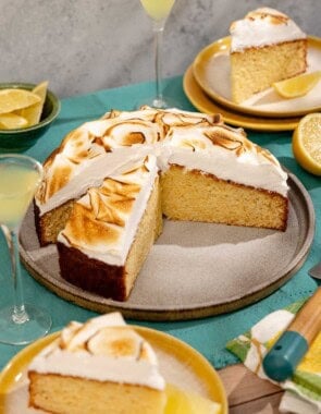 A limoncello cake on a platter with a piece cut from it. Next to this is a bowl of lemon wedges, two plates each with a piece of the cake, 2 glasses of limoncello, and a lemon half.