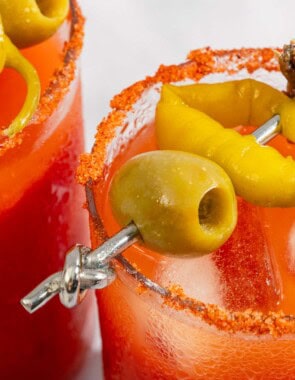 A close up of two Mediterranean bloody Marys garnished skewers of olives, pickled peppers and anchovies.