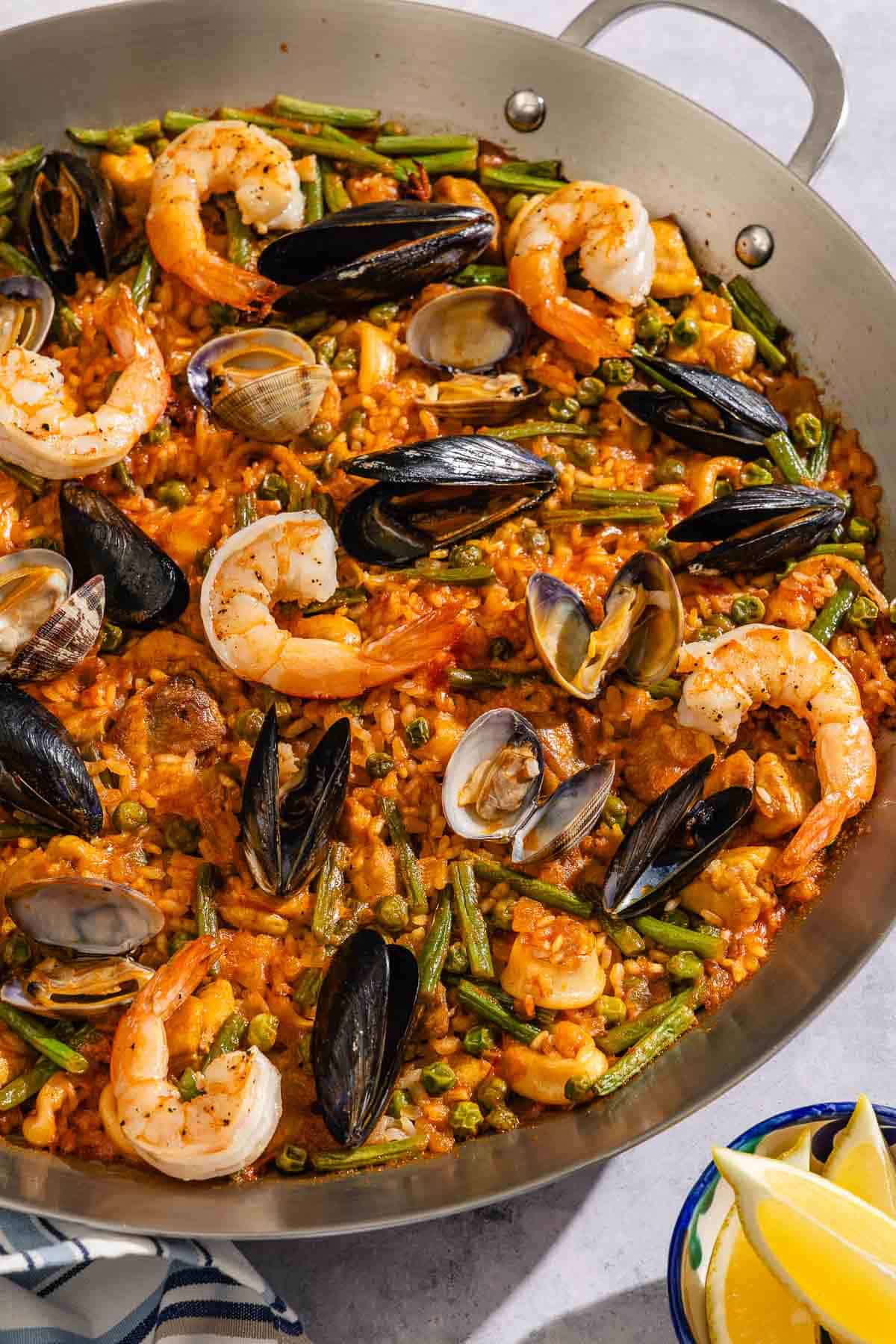 Paella in a paella pan next to a kitchen towel and a bowl of lemon wedges.