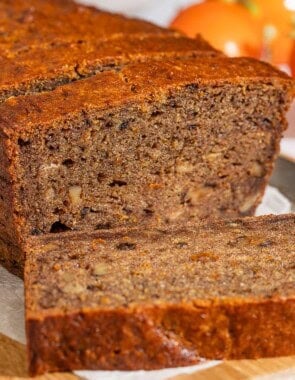 A partially sliced loaf of persimmon bread behind 3 slices of the bread on a parchment-lined cutting board with a knife. In the background are 3 persimmons.