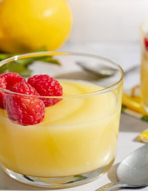 Lemon pudding topped with raspberries in a small glass bowl next to a spoon, another bowl of pudding, and several lemons.
