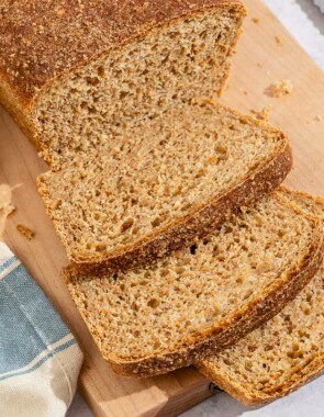 A partially sliced whole wheat bread loaf behind 3 slices of the bread on a cutting board. Next to this is a kitchen towel and a knife.