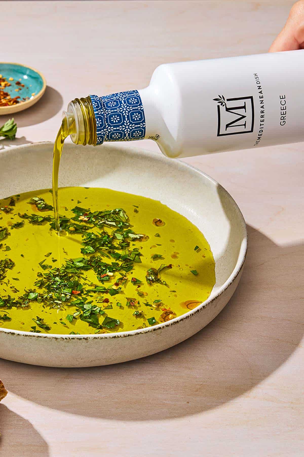 Olive oil being poured from a bottle into a bowl of olive oil seasoned with spices and herbs. Next to this is a small bowl of red pepper flakes.