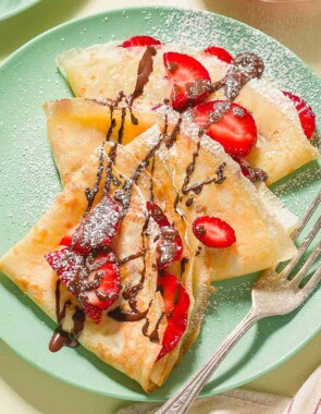 Three strawberry crepes drizzled with chocolate on a plate with a fork.