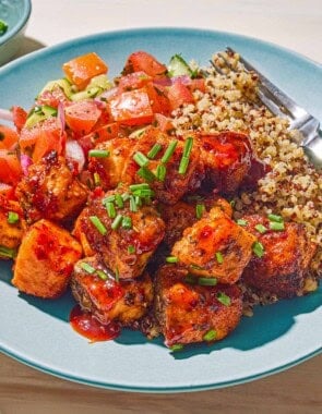 A serving of the air fryer salmon bites topped with chopped chives on a plate with quinoa, tomato cucumber salad and a fork.