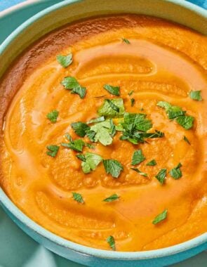 A bowl of red lentil soup topped with parsley on a plate with a spoon and a piece of crusty bread.