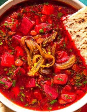 A bowl of beet soup topped with fried onions and a piece of pita bread next to more pita bread.