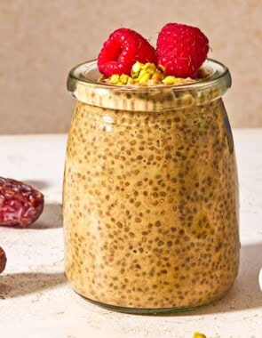 A jar of chia seed pudding topped with raspberries and crushed pistachios next to three whole dates and a bowl of crushed pistachios.