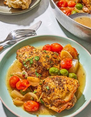 A serving of the chicken provencal on a plate. Next to this is a knife, a fork, a plate with French baguette pieces and a skillet with the rest of the recipe and a serving spoon.