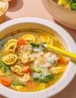 Chicken tortellini soup topped with grated Parmigiano Reggiano cheese in a bowl with a spoon. Next to this is a large pot with the rest of the soup and bowls of pepper and grated Parmigiano Reggiano cheese.