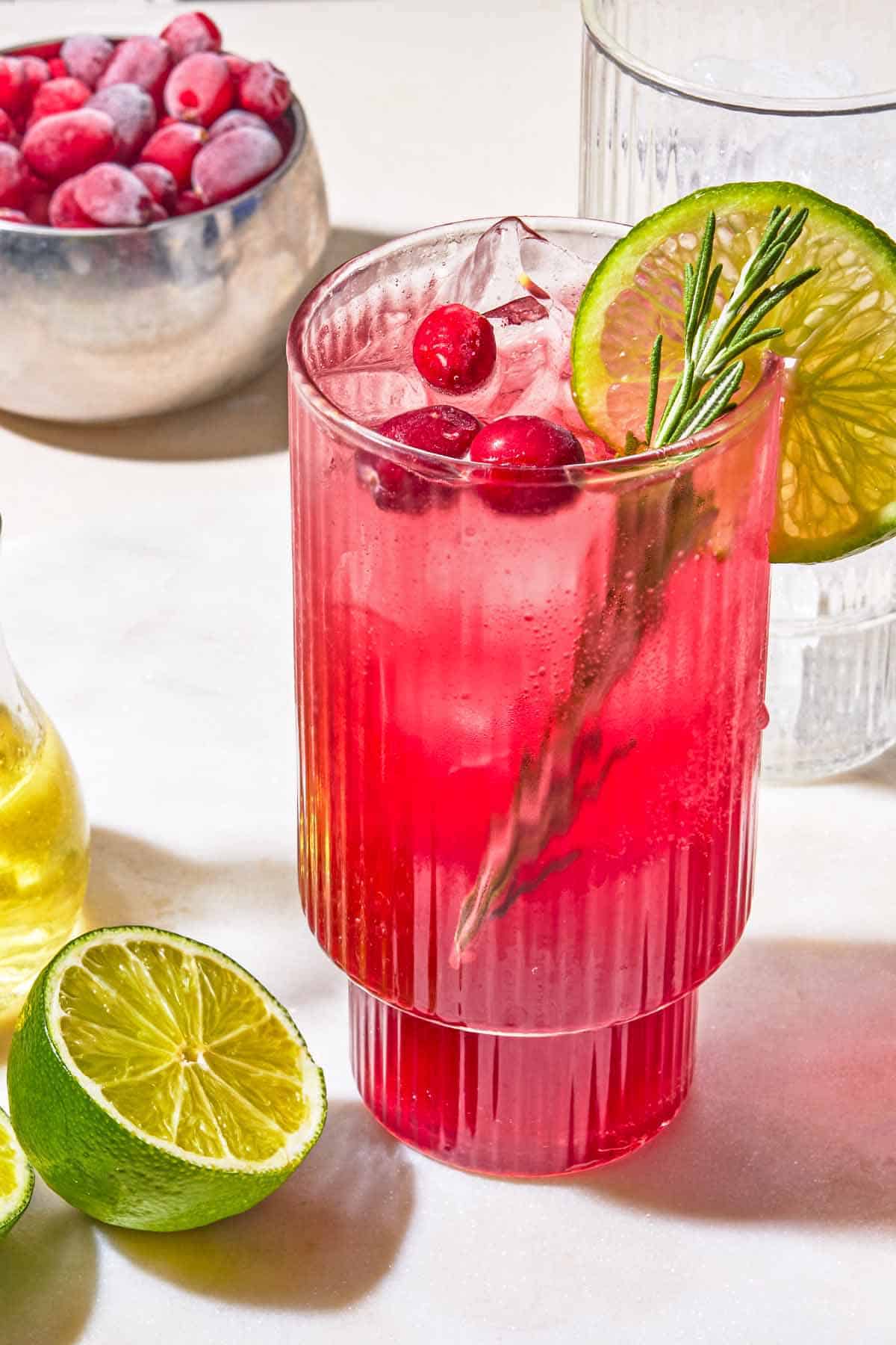 A glass of cranberry mocktail garnished with rosemary, cranberries and lime next to a lime half, an empty glass and a bowl of cranberries.