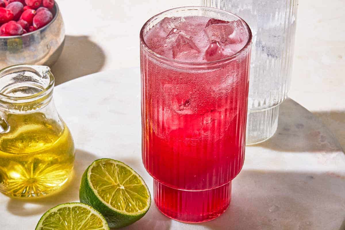 A glass of cranberry mocktail next to limes, simple syrup I a bottle, a bowl of cranberries and a glass of ice.