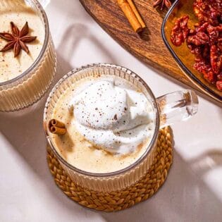 Two mugs of dairy-free eggnog garnished with star anise, a cinnamon stick and whipped coconut cream next to a wooden tray with a bowl of dates, a cinnamon stick a star anise.
