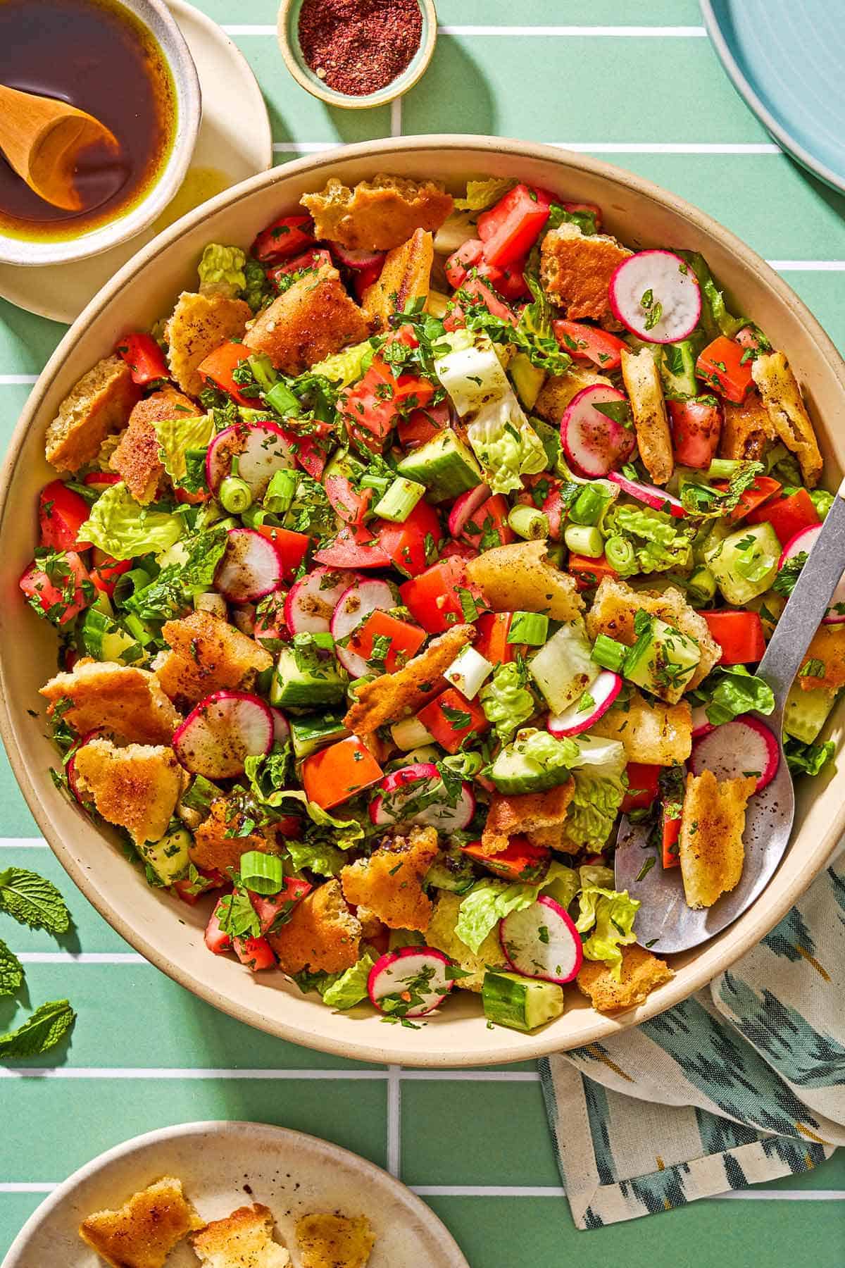 Fattoush in a serving bowl with a spoon. Next to this is a small bowl of the dressing and sumac, a cloth napkin, and a plate of toasted pita pieces.