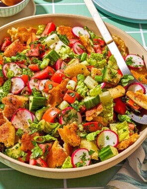Fattoush in a serving bowl with a spoon. Next to this is a small bowl of the dressing and sumac, a cloth napkin, and a plate.
