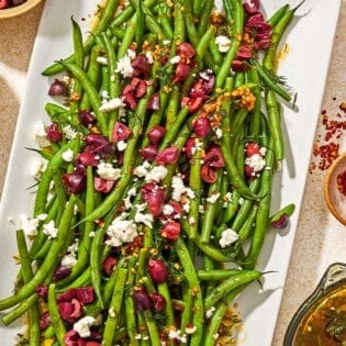 Green bean salad on a platter next to bowls of feta and red pepper flakes, and the dressing for the salad in a measuring cup.