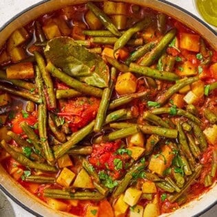 Fasolakia in a large pot next to a small bowl of chopped parsley, a juiced lemon half and a bottle of olive oil.