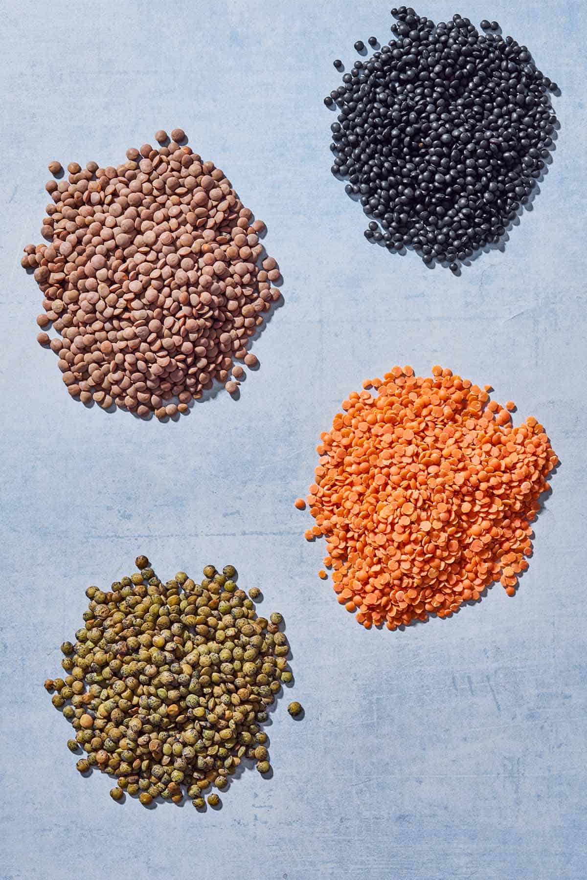 4 piles of lentils including red lentils, green lentils, black lentils and brown lentils.