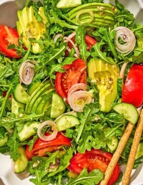 Arugula salad with tomato, cucumber and avocado in a serving bowl with serving utensils next to a bowl of the dressing.