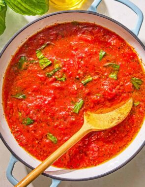 Marinara sauce in a skillet with a wooden spoon. Next to this is a bottle of olive oil and sprigs of fresh basil.