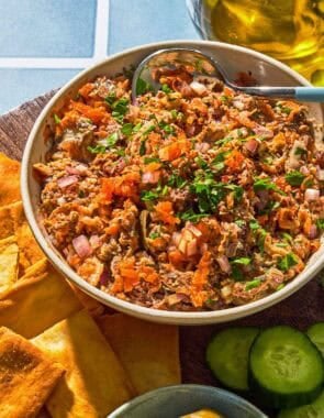 Sardine dip in a bowl with a spoon next to cucumber slices, pita chips, a bowl of olives and a bottle of olive oil.