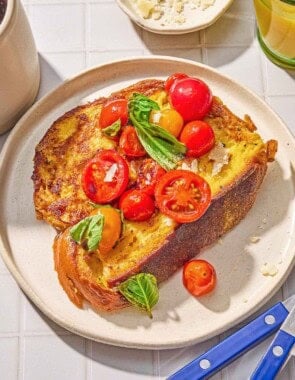 A slice of the savory french toast with the veggie topping on a plate. Next to this is a knife and fork, a small bowl Parmigiano Reggiano cheese, and a cup of orange juice.