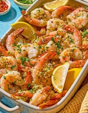 Shrimp oreganata with lemon wedges in a baking dish next to bowls of chopped parsley and red pepper flakes.