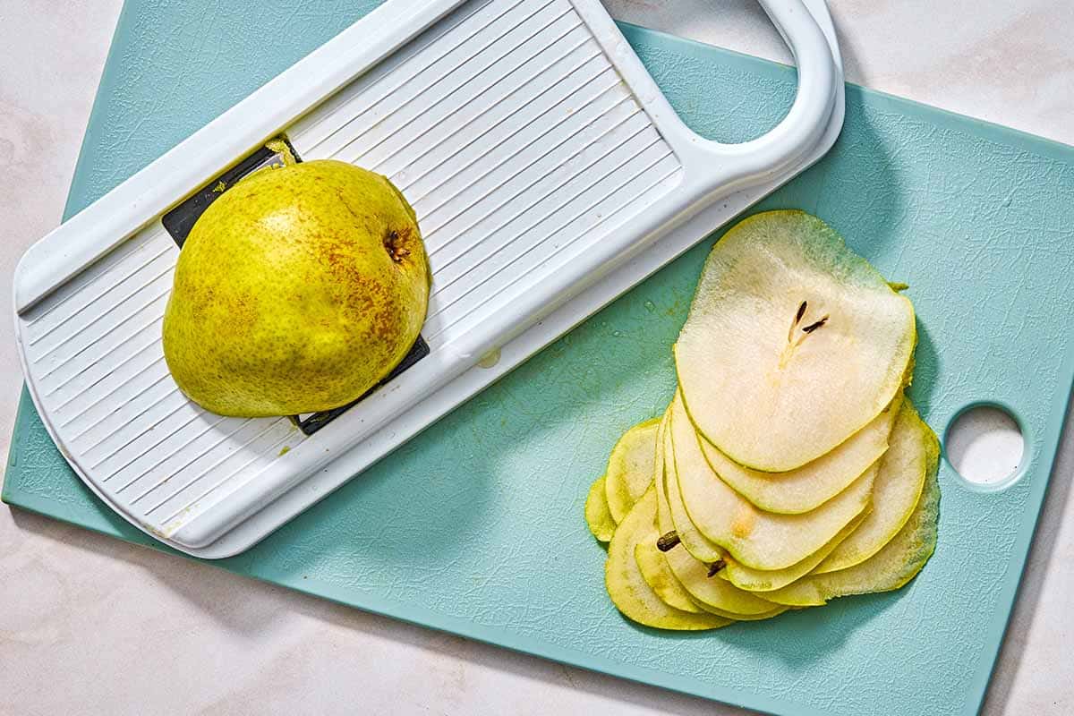 A mandolin slicer holding a pear half on a cutting board with thinly sliced pieces of the pear.