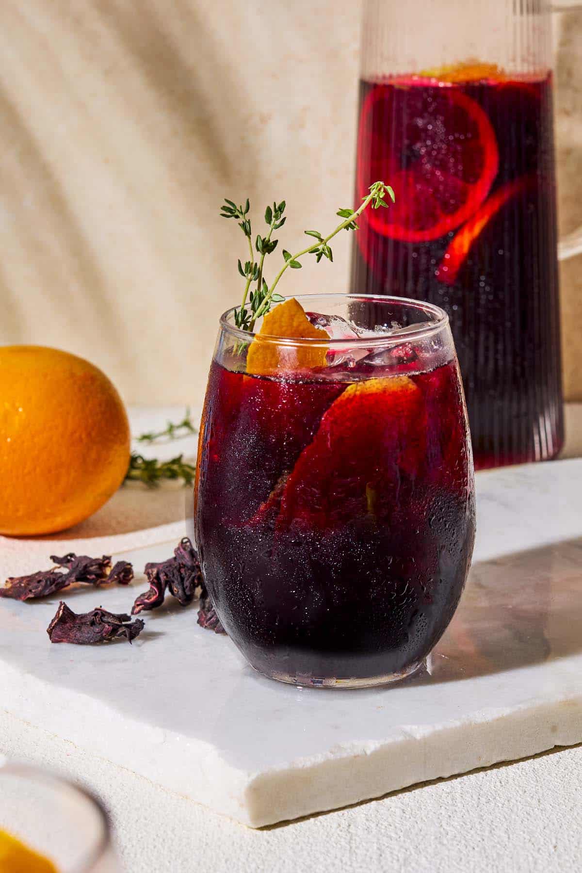 A close up of a glass of non alcoholic sangria garnished with sprigs of fresh thyme and orange and lemon peels on a marble serving tray. Behind this is a pitcher of the sangria, dried hibiscus flowers, and an orange.