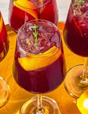 Four tart cherry juice mocktails garnished with lemon twists and sprigs of fresh thyme on a tray with a lemon.