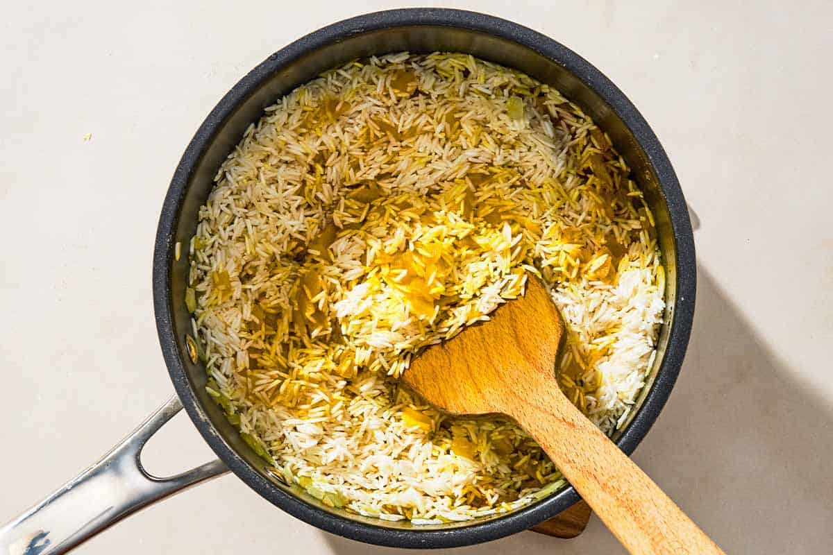 The aromatics and rice for the turmeric rice being stirred together in a saucepan with a wooden spoon.