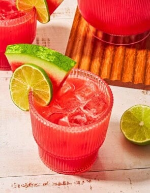 A glass of watermelon sangria garnished with a lime wheel and a watermelon slice next to another glass of the sangria, a pitcher of the sangria, and a lime half.